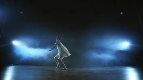 The-zoom-camera-moves-the-woman-dancer-across-the-stage-with-software-and-smoke.-A-modern-dramatic-ballet-a-woman-in-a-white-dress-spins-on-one-leg-and-jumps..